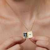 A person is holding a gold necklace with a small square pendant that contains a photograph of two people.