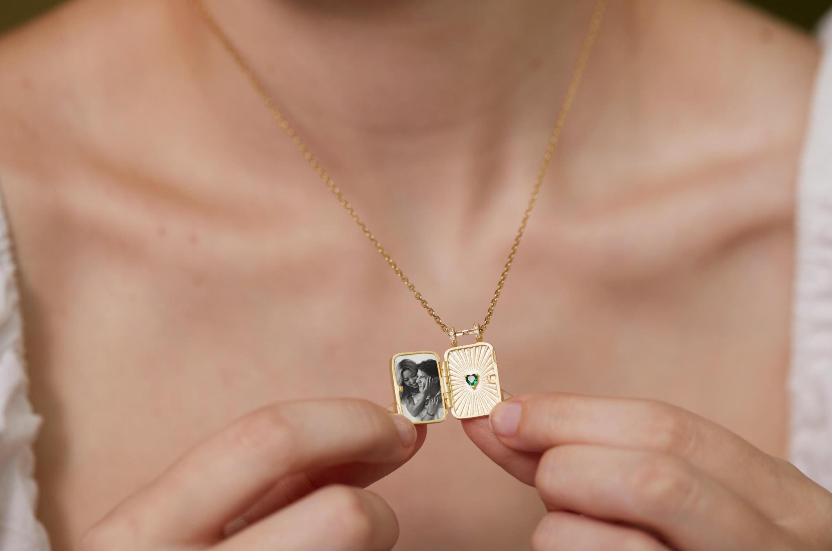 A person is holding a gold necklace with a small square pendant that contains a photograph of two people.