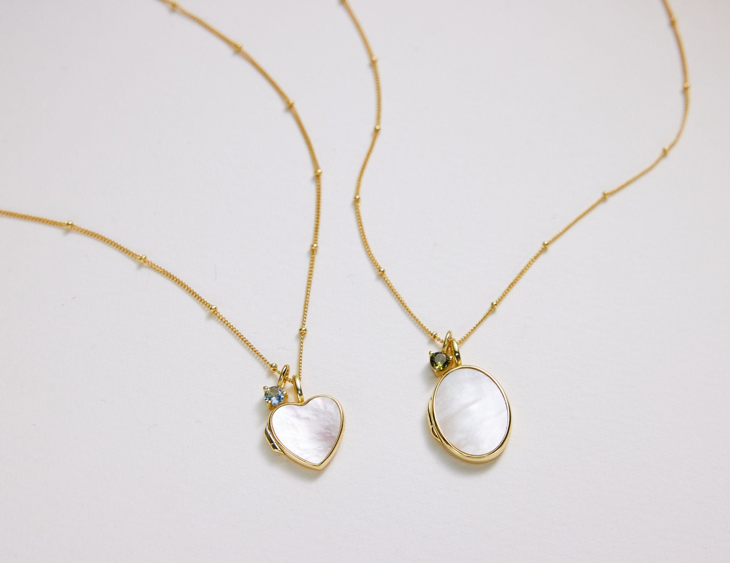 Two gold necklaces with heart and oval pendants on a light gray background