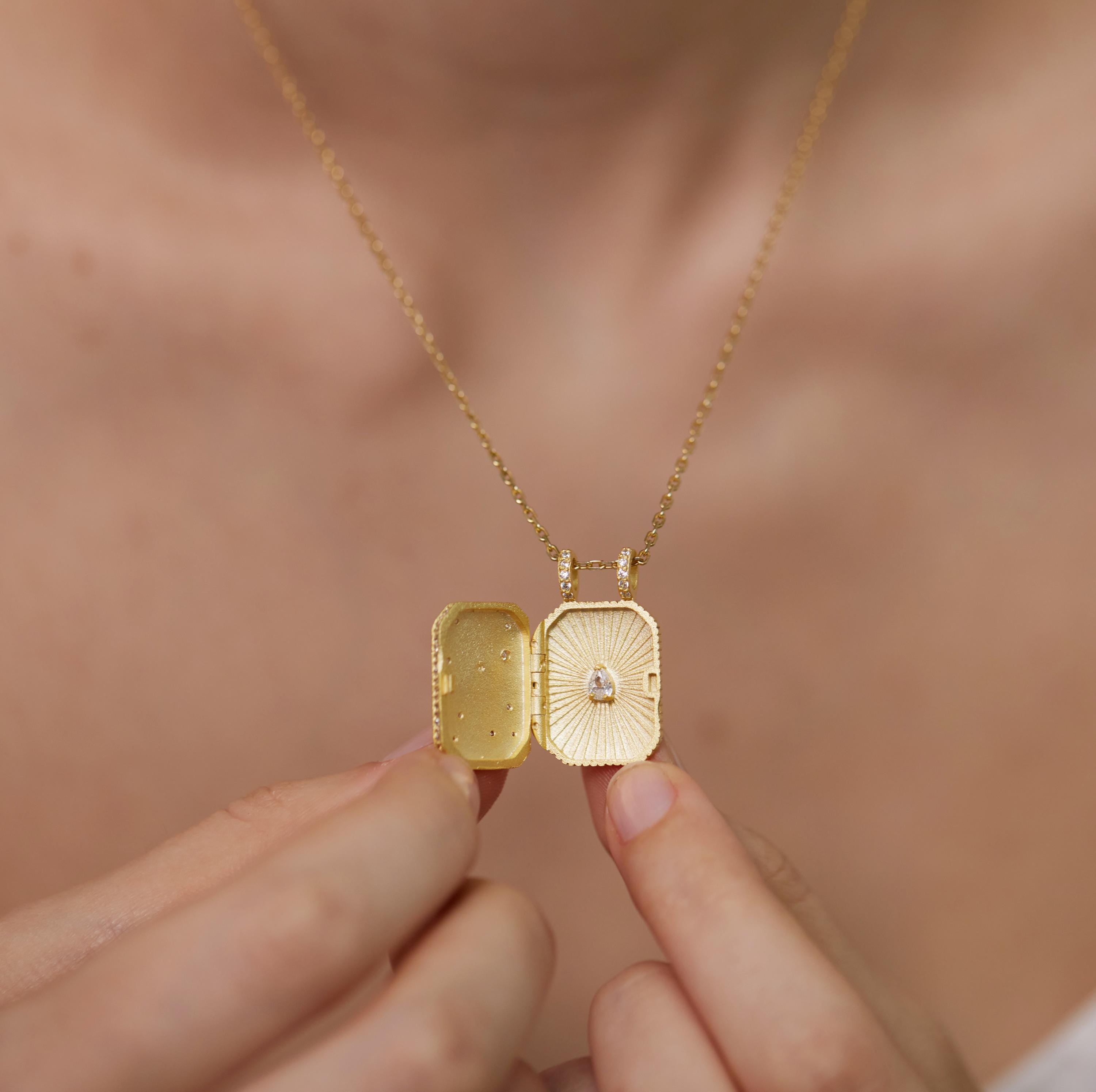 A person's hand holding a gold-colored necklace with an open pendant, revealing a small diamond.