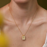 A gold necklace with a square pendant is worn around a person's neck.