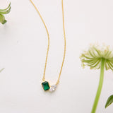 A gold necklace with a green gemstone pendant is displayed on a white surface, surrounded by small white flowers.
