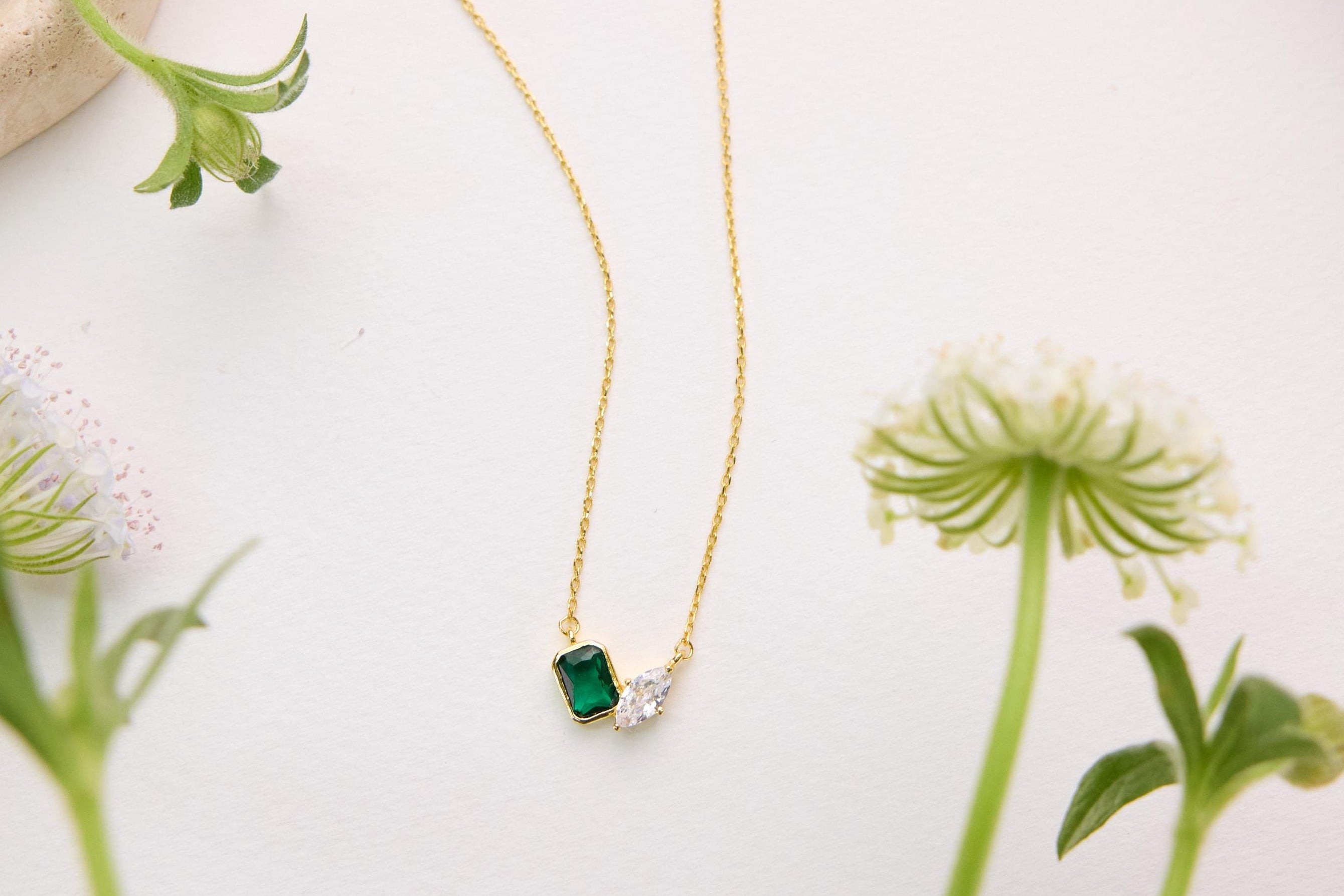 A gold necklace with a green gemstone pendant is displayed on a white surface, surrounded by small white flowers.