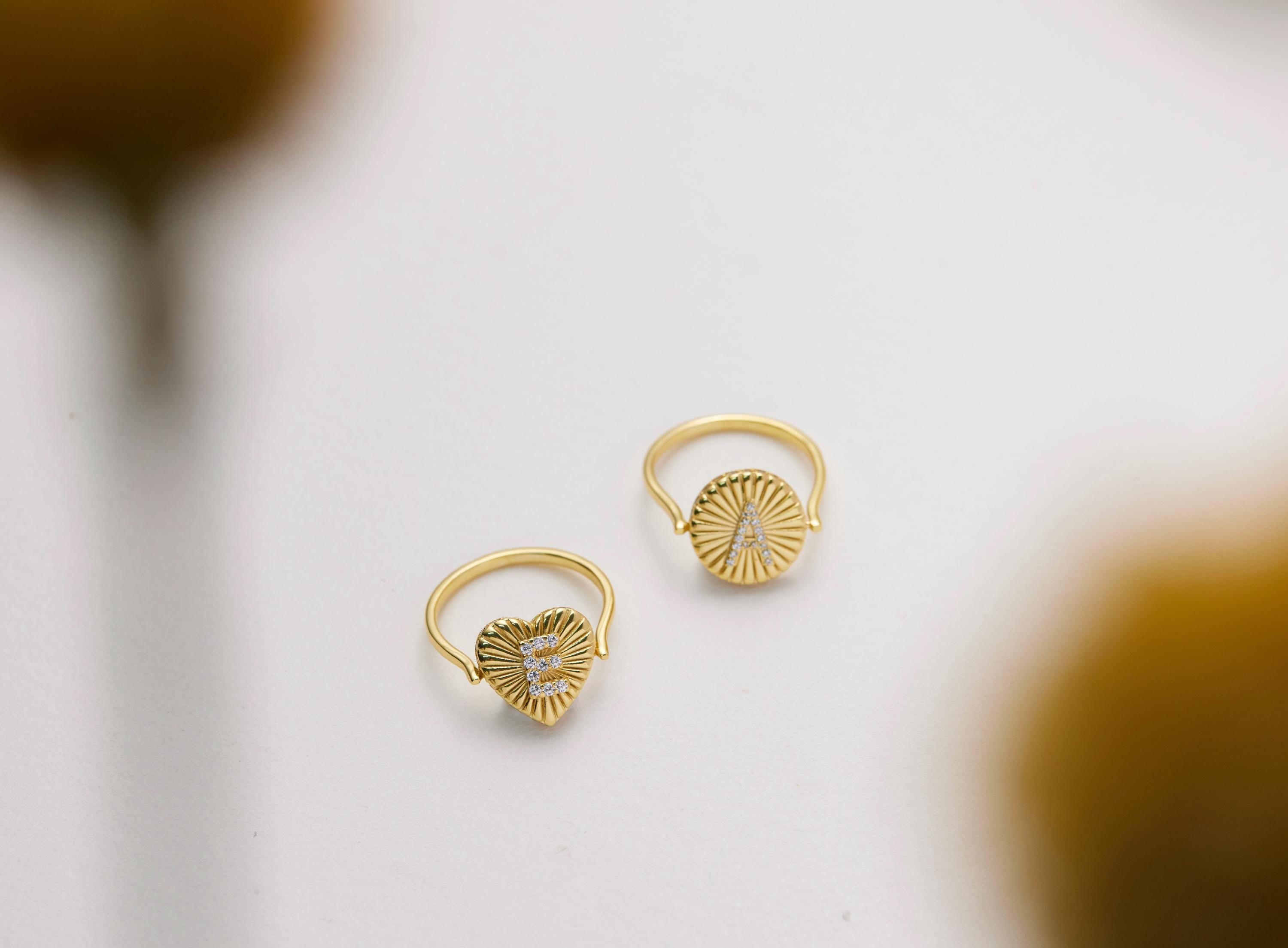 Two gold rings with heart-shaped designs and letters on them are placed on a white surface.
