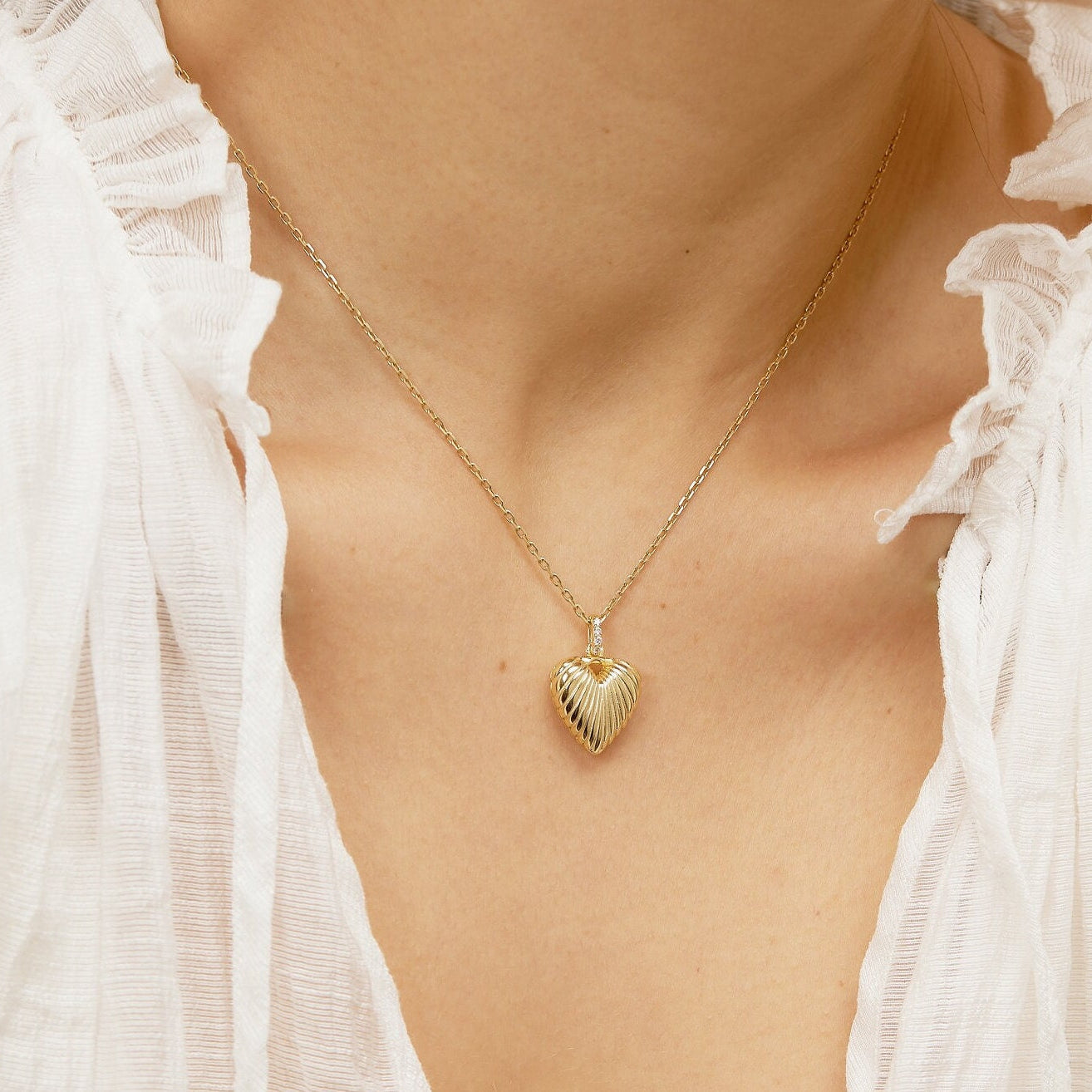 Gold necklace with a heart-shaped pendant worn by a person in a white outfit.