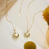 two gold heart-shaped locket charms with engraved initials, displayed on a white surface alongside a yellow flower and a glass vase.