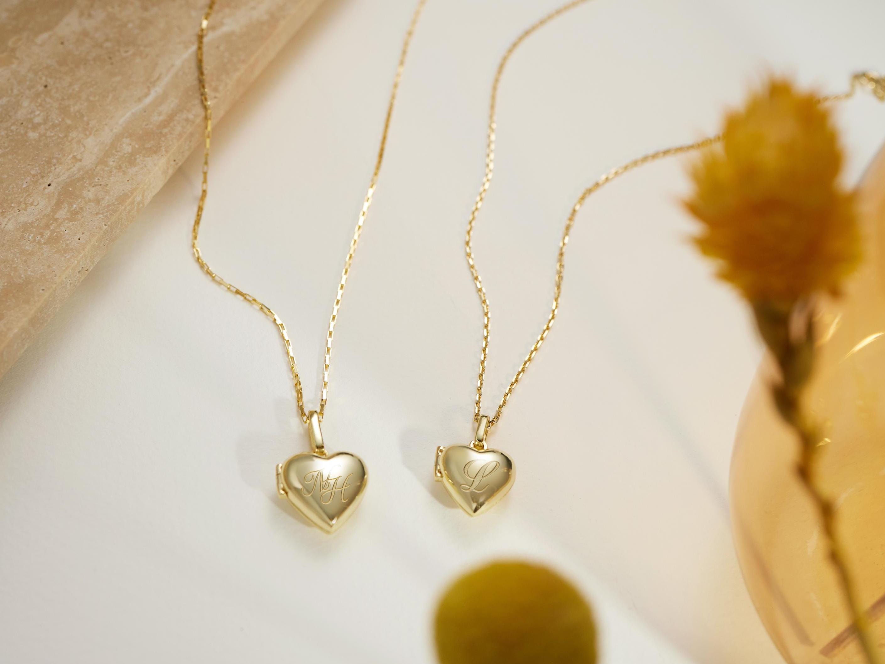 two gold heart-shaped locket charms with engraved initials, displayed on a white surface alongside a yellow flower and a glass vase.