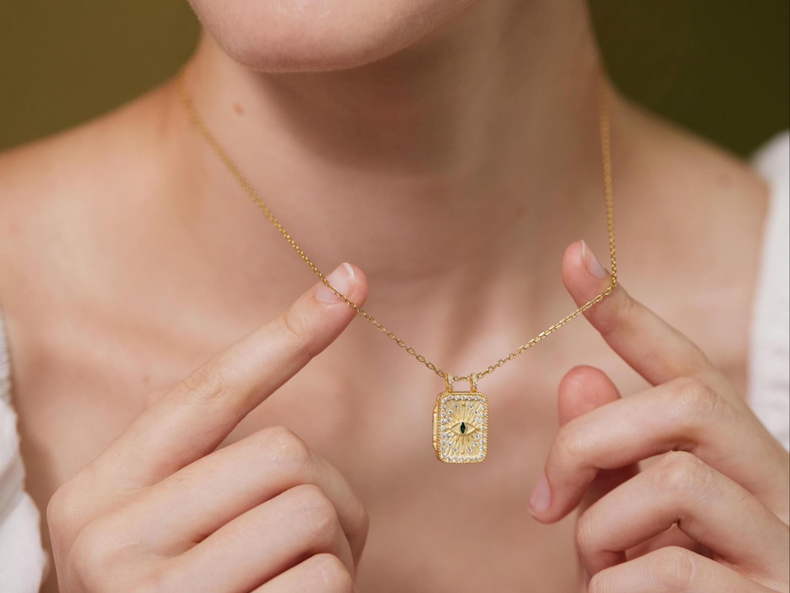 A close-up of a person&#39;s hand holding a gold necklace with a pendant, wearing a white blouse.