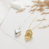 Two heart-shaped necklaces, one gold and one silver, on a light background.