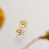 Two gold rings with heart-shaped designs are displayed against a white background, with a blurred yellow flower in the foreground.