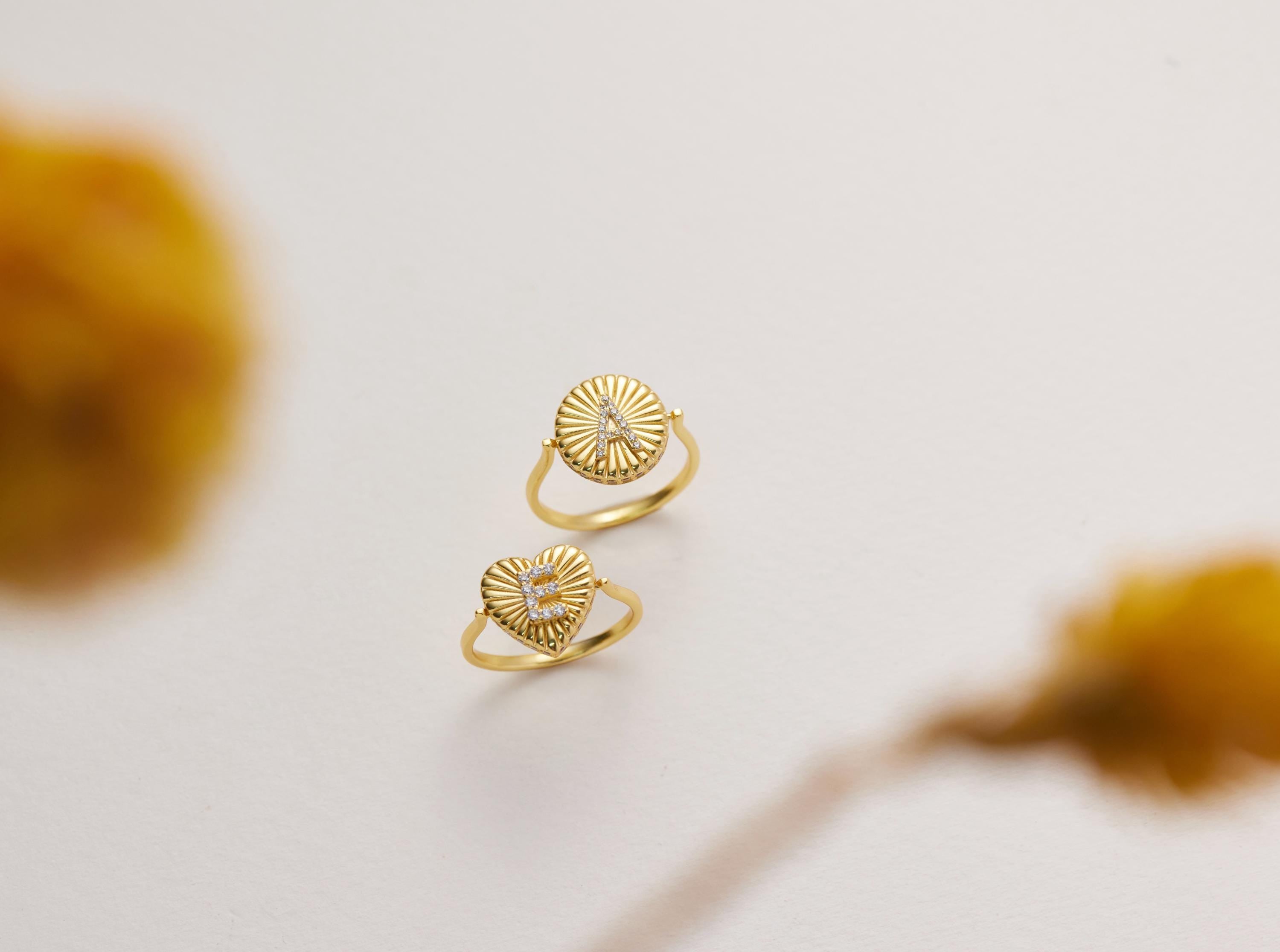 Two gold rings with heart-shaped designs are displayed against a white background, with a blurred yellow flower in the foreground.