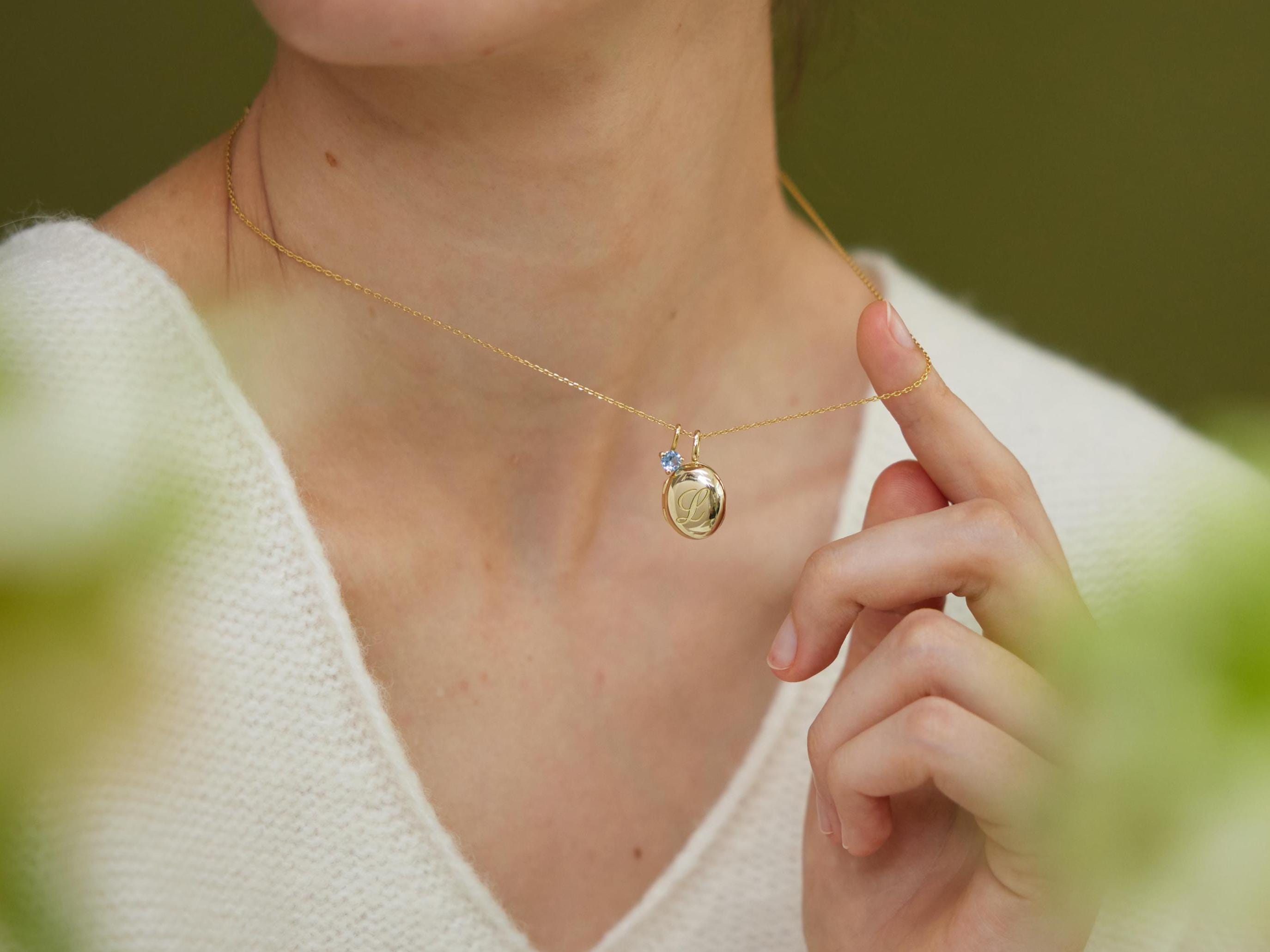 Close-up of a person wearing a gold necklace with a pendant, with a blurred green background.