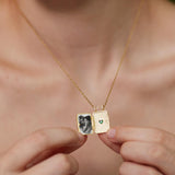 A person is holding a gold necklace with a small square pendant that contains a photograph of two people.