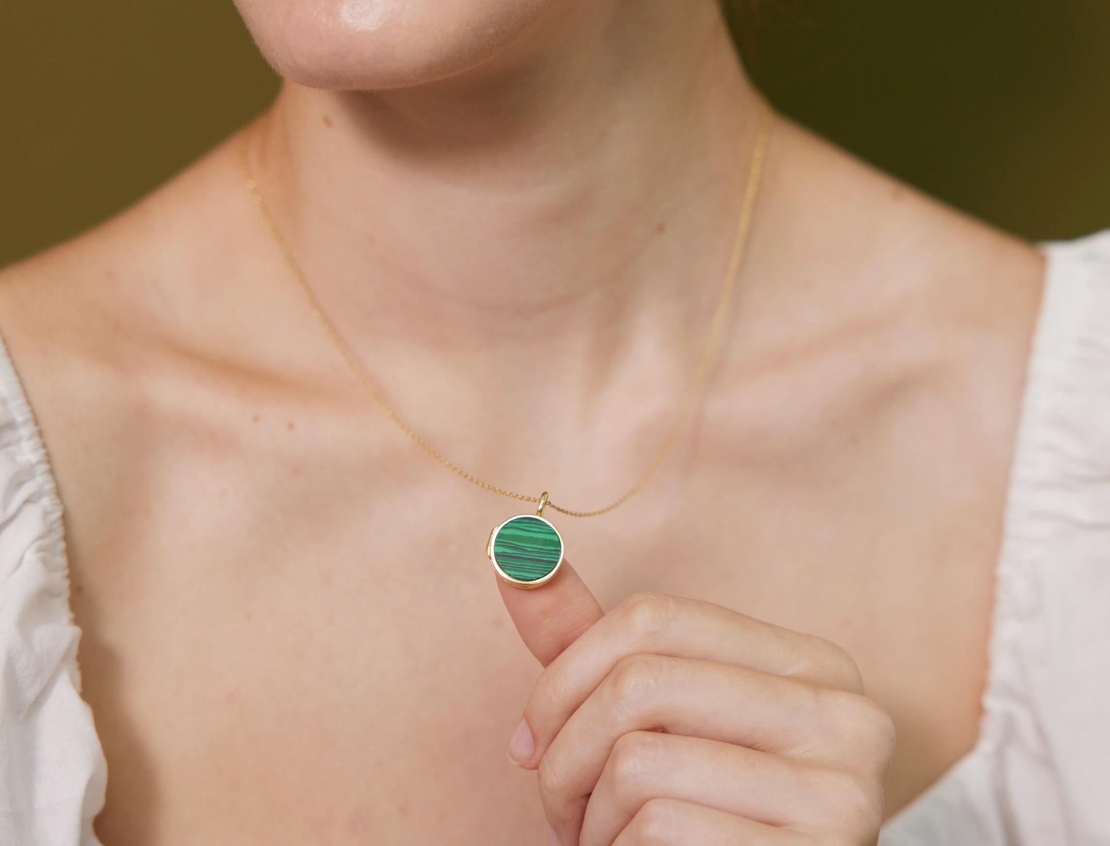 A close-up of a person's neck and chest area, with a gold necklace and a green pendant visible. The person is holding the pendant up to their face, possibly to show it off or to admire it.