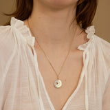 Gold necklace with a circular pendant worn by a person in a light blouse against a neutral background