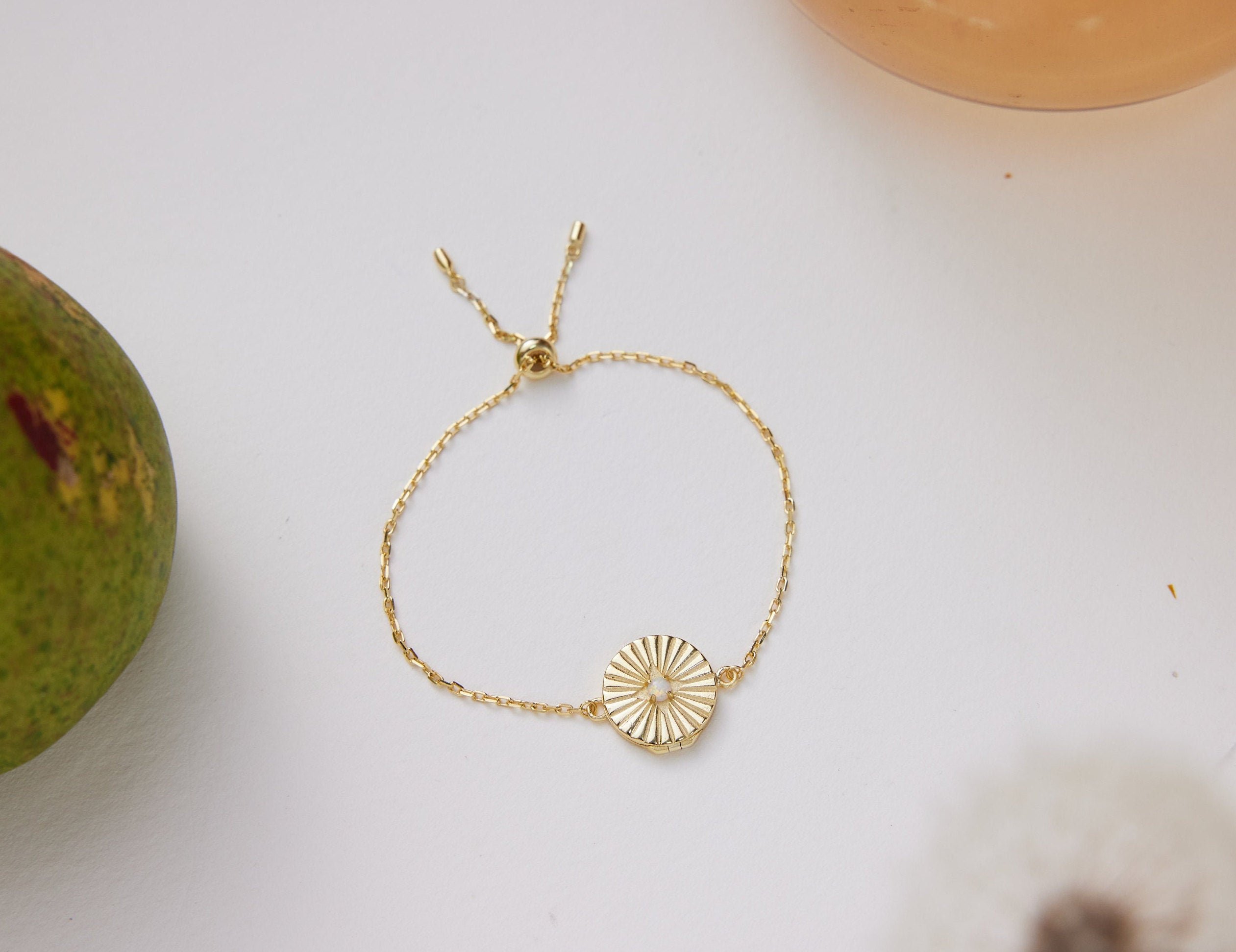 Gold bracelet with a circular design on a white surface next to a green fruit.