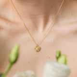 Gold necklace with a floral pendant on a person's neck, with green flowers in the background.