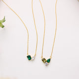 two gold necklaces with gemstones, including green and white stones, hanging on a white surface.