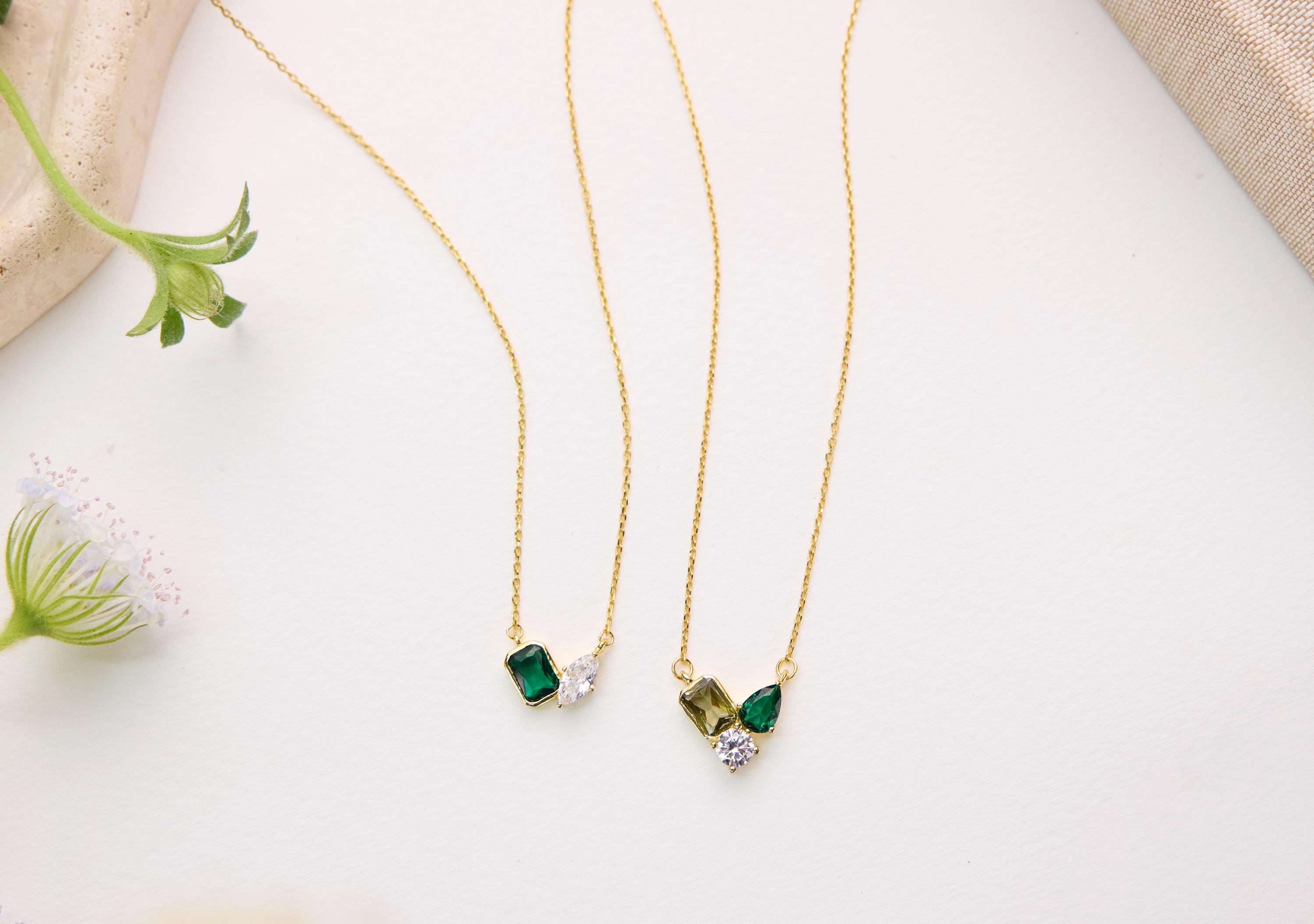 two gold necklaces with gemstones, including green and white stones, hanging on a white surface.