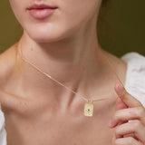 A close-up of a woman wearing a gold necklace with a pendant, posing against a green background.