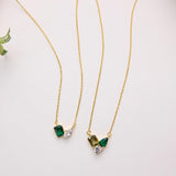 two gold necklaces with green and clear gemstone pendants, placed on a white surface alongside a small green flower.