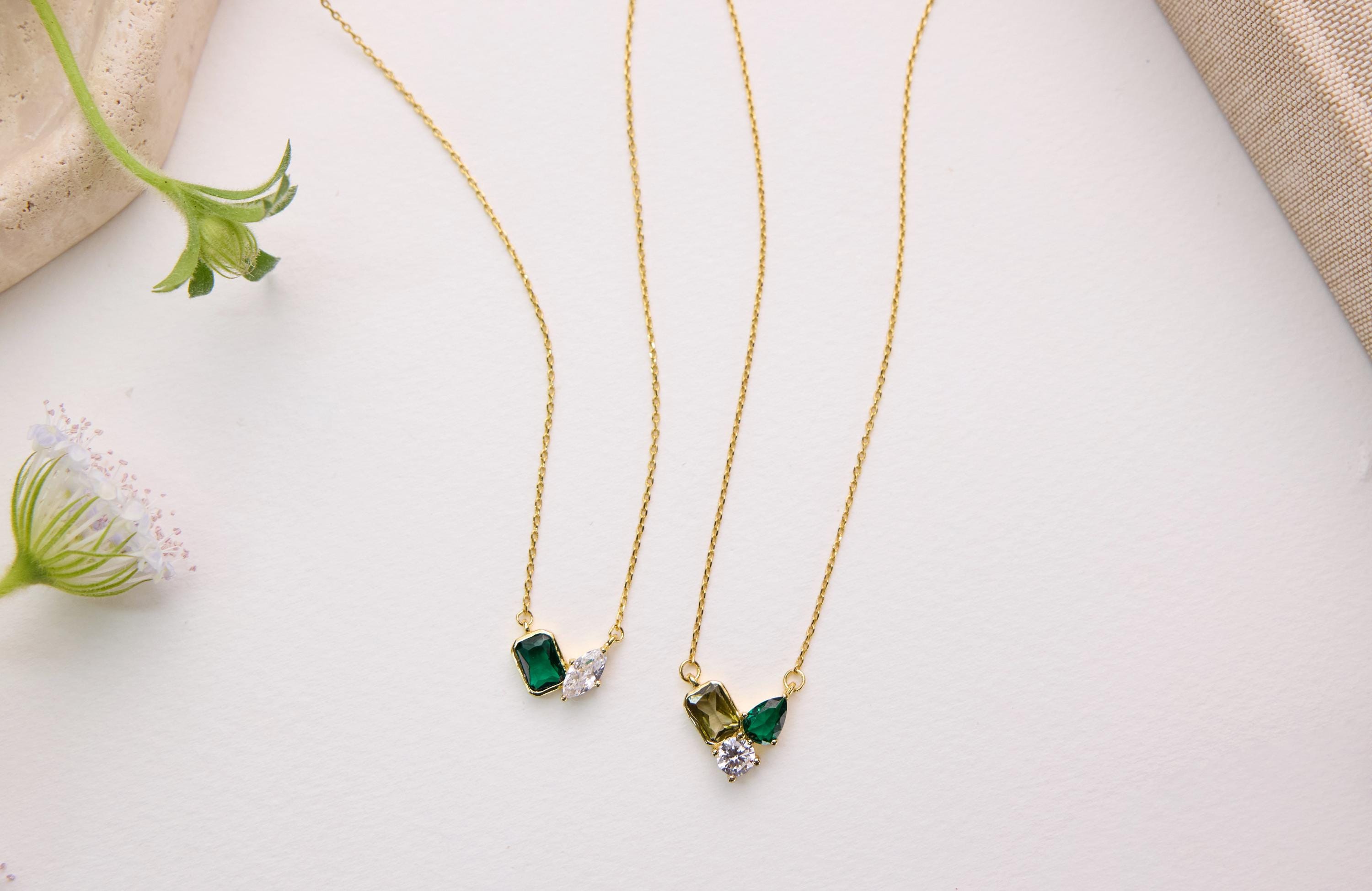 two gold necklaces with green and clear gemstone pendants, placed on a white surface alongside a small green flower.