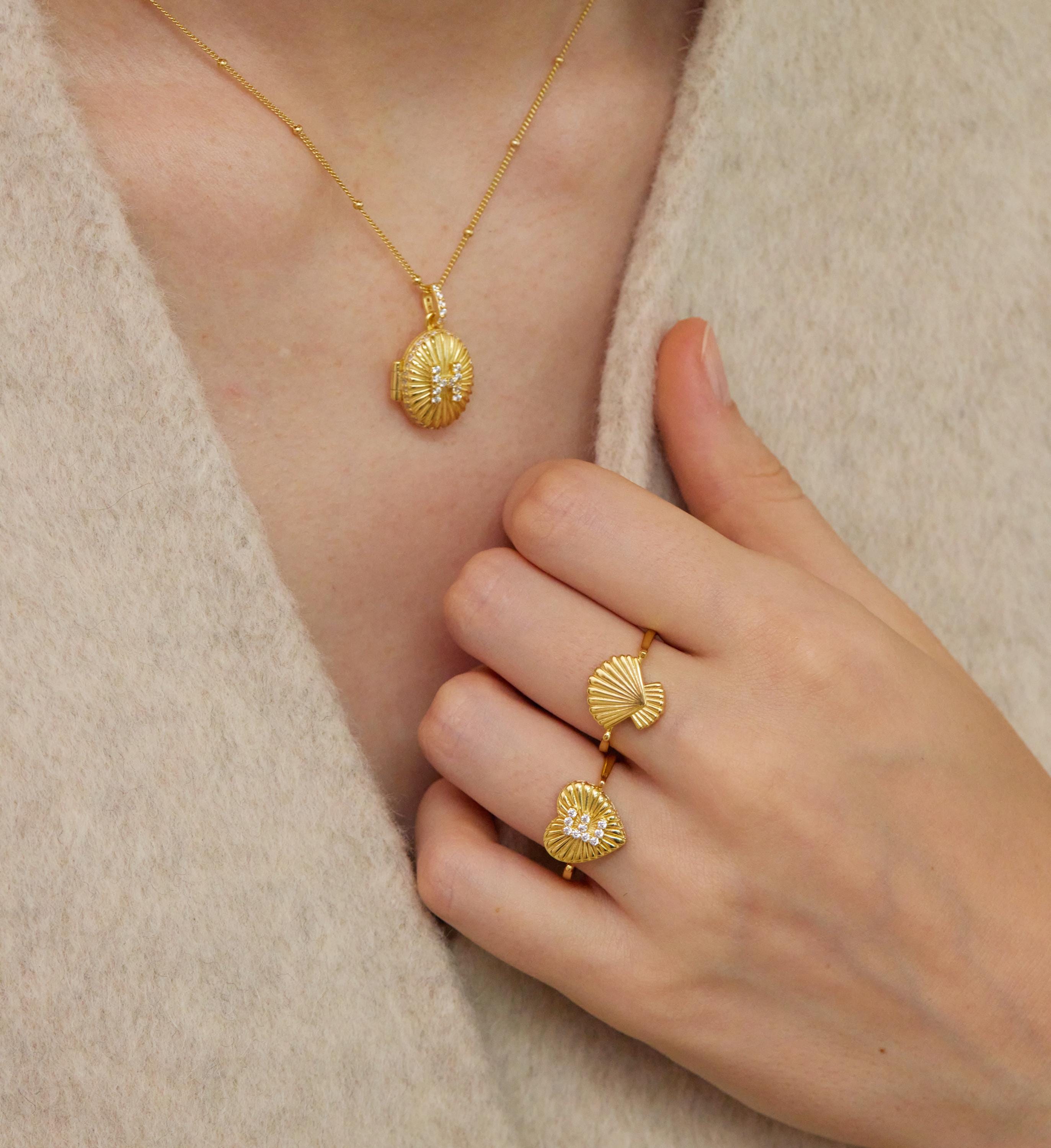 A close-up of a person's hand wearing three gold-colored rings with unique designs, including a ring with a shell-like pattern and another ring with a heart-shaped design.