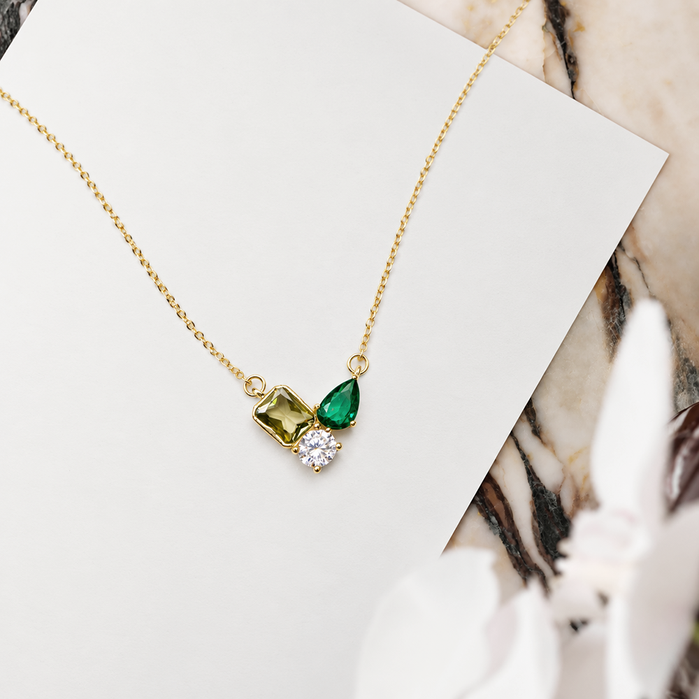 A delicate gold chain necklace with a small pendant featuring a green rectangular gemstone, a green teardrop gemstone, and a clear round crystal cluster, displayed on a white card over a marble surface.