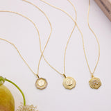 Three gold-colored necklaces with pendants are displayed on a white surface, accompanied by a pear and some flowers.
