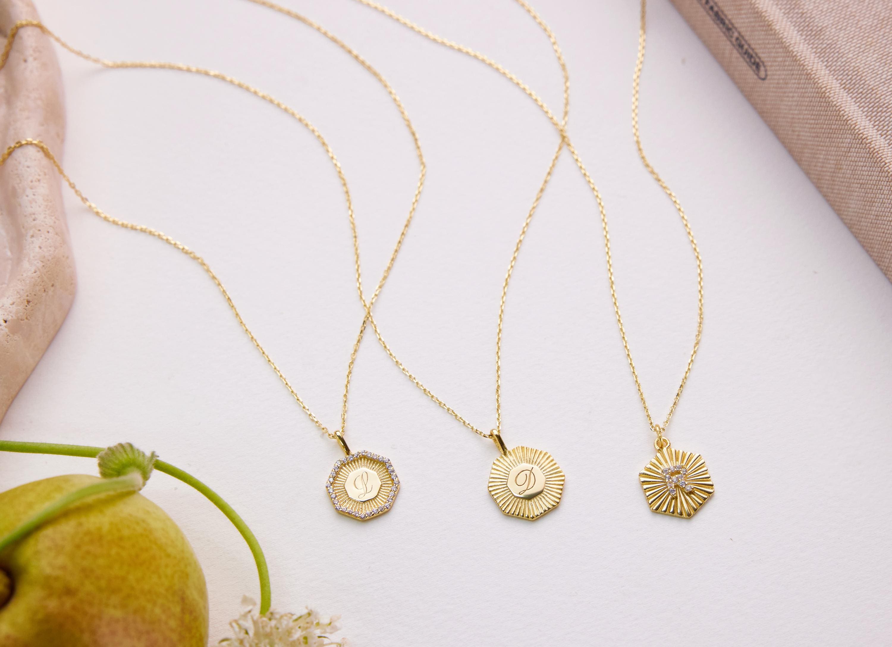 Three gold-colored necklaces with pendants are displayed on a white surface, accompanied by a pear and some flowers.