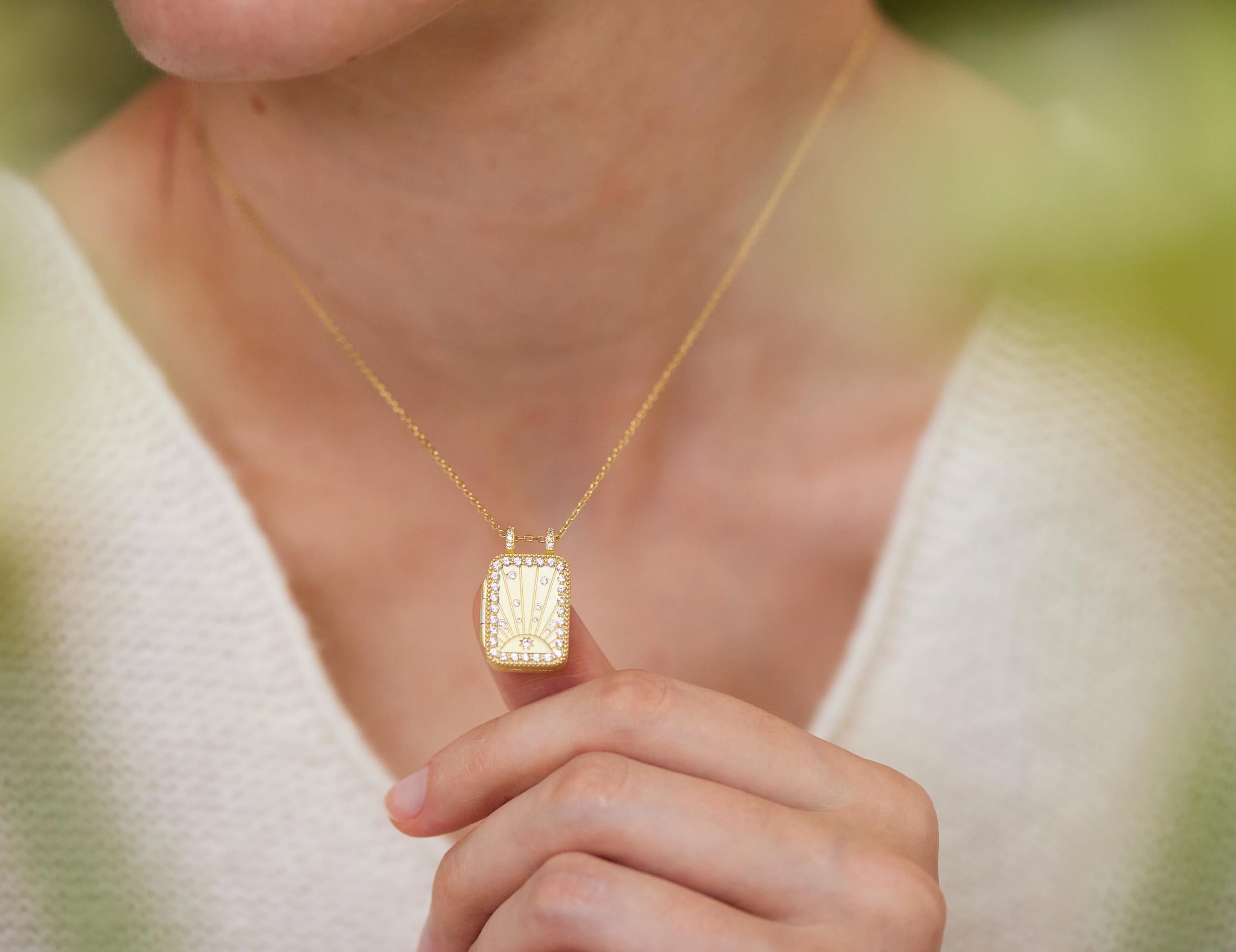 A person wearing a gold necklace with a pendant is holding the pendant up to their face.
