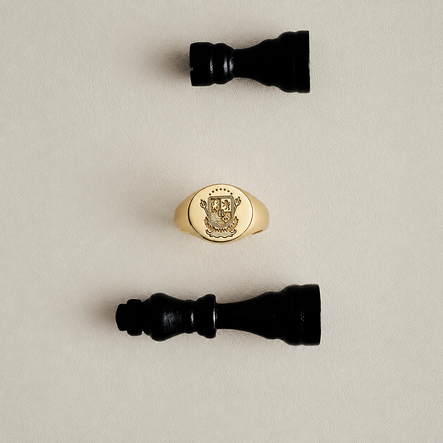 A gold signet ring with an engraved crest is displayed vertically in the center, flanked by two black chess pieces on a light beige textured background.