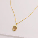A delicate gold necklace with a small oval-shaped pendant is displayed against a plain white background.