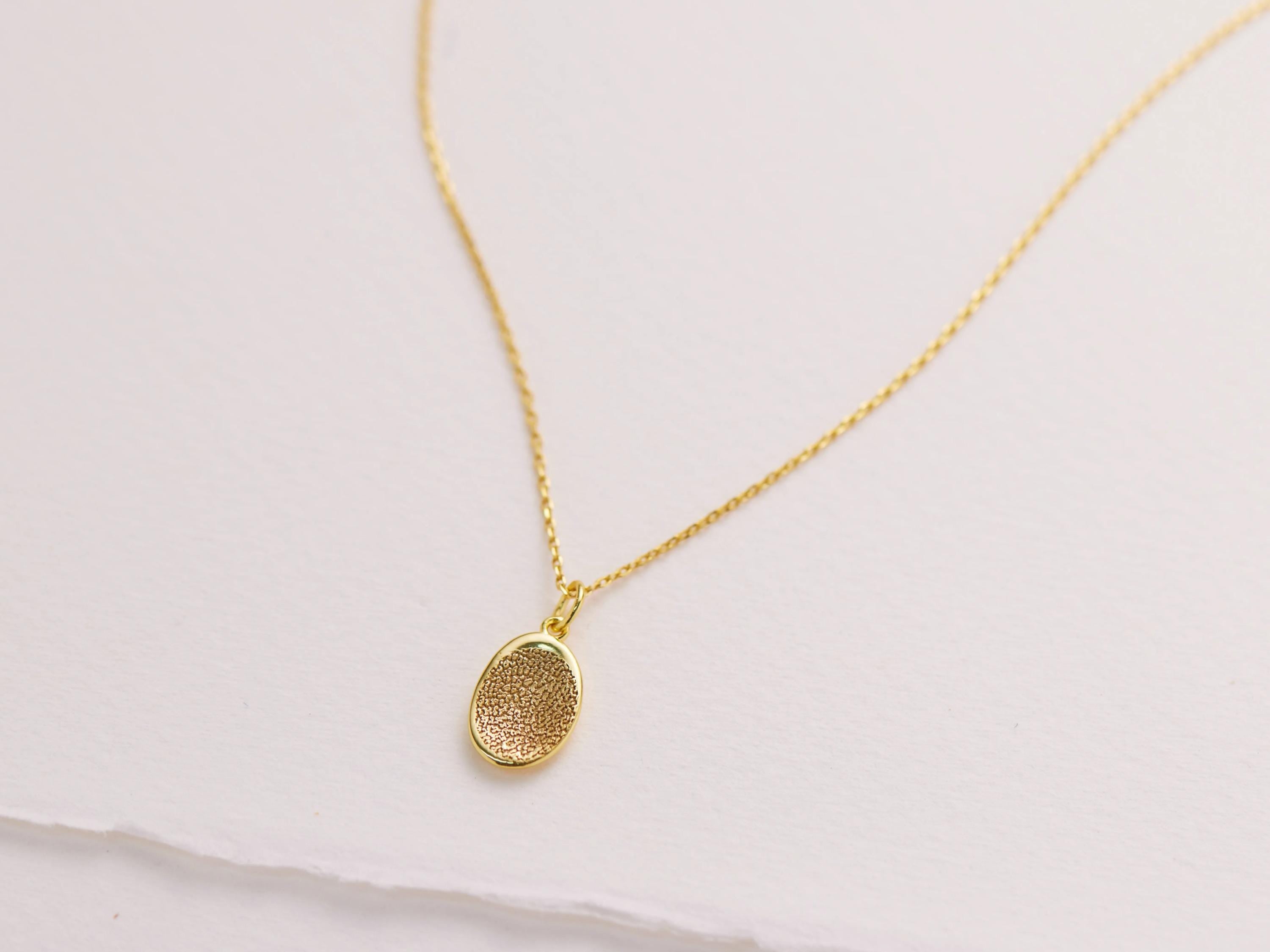 A delicate gold necklace with a small oval-shaped pendant is displayed against a plain white background.