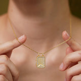 A person is holding a gold necklace with a rectangular pendant featuring a floral design.
