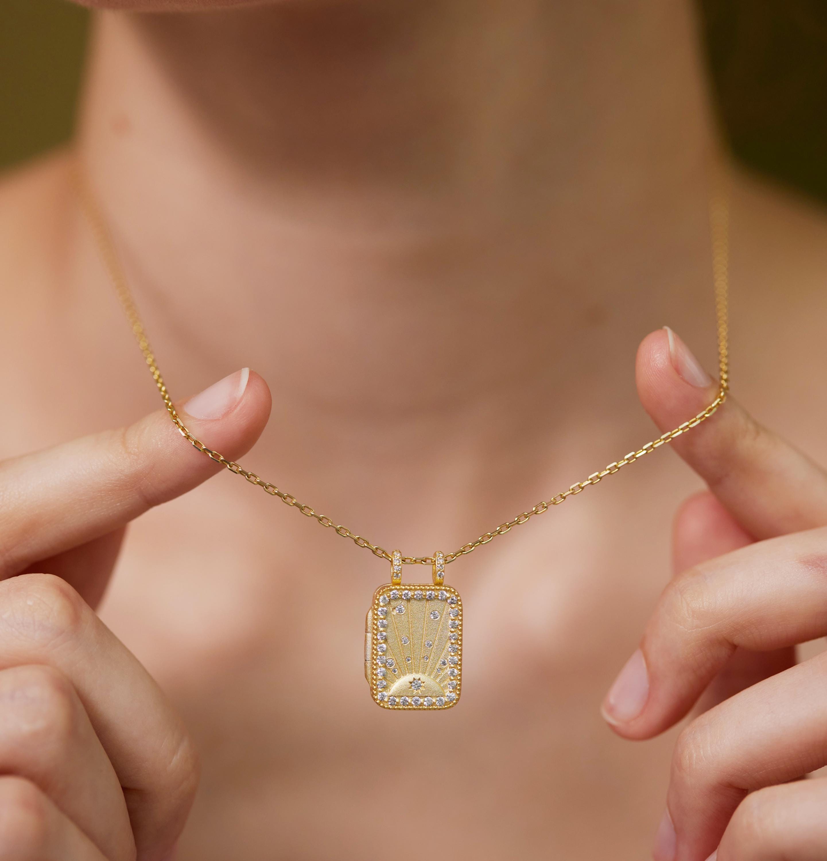 A person is holding a gold necklace with a rectangular pendant featuring a floral design.