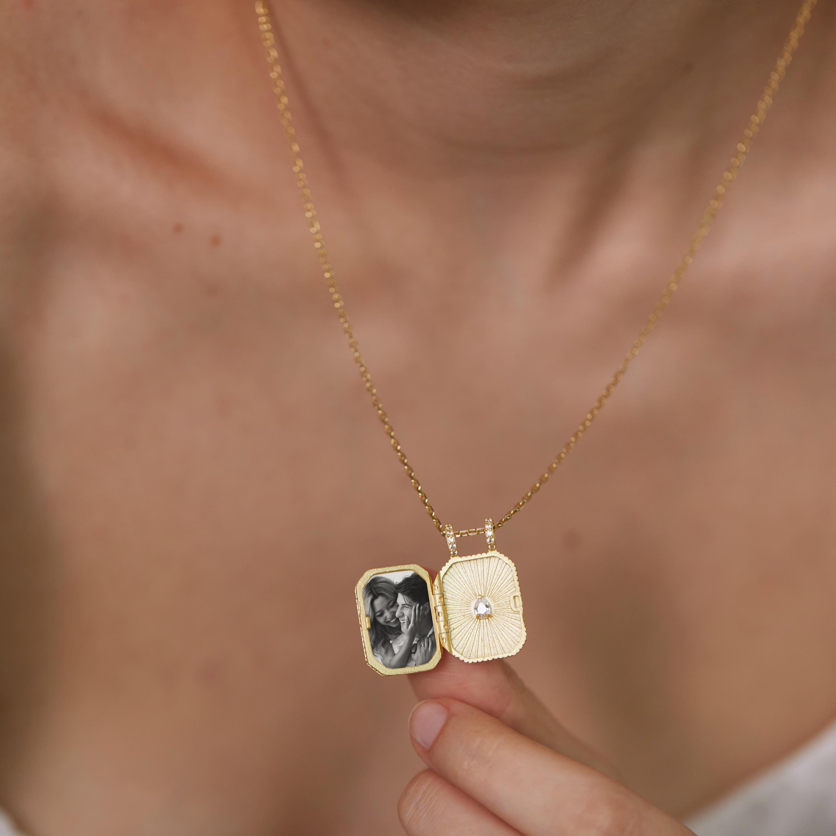 A person is holding a gold-colored necklace with a small, rectangular pendant that contains a black and white photograph.