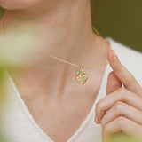 A person wearing a white sweater is holding a gold heart-shaped pendant necklace with a small charm on it.