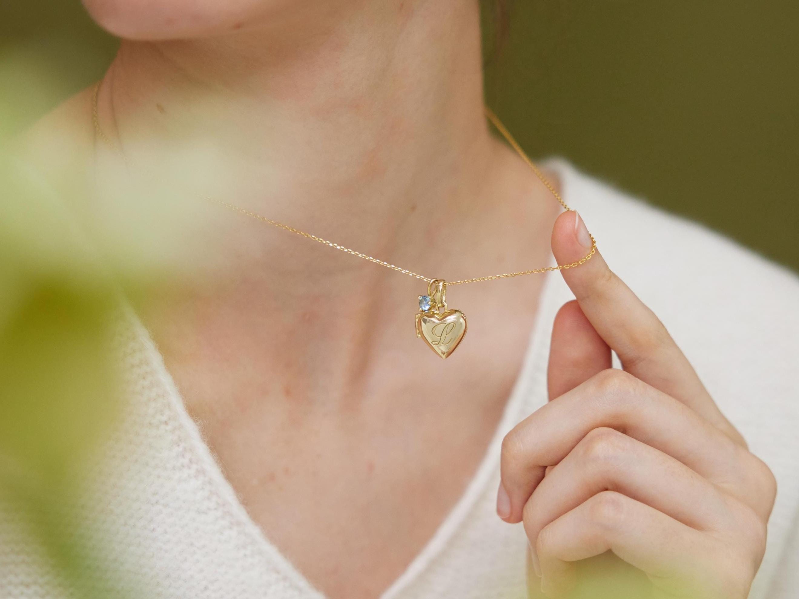 A person wearing a white sweater is holding a gold heart-shaped pendant necklace with a small charm on it.