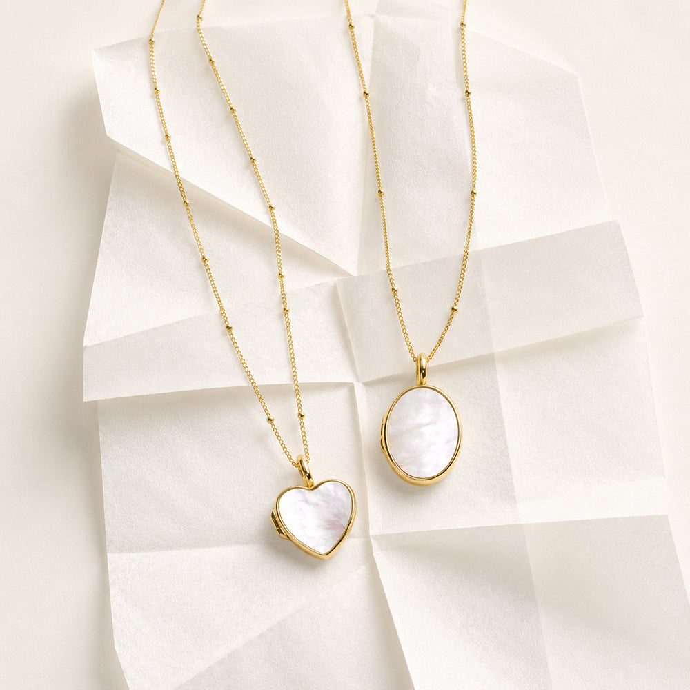 Two gold pendant necklaces displayed on white tissue paper. One necklace has a heart-shaped pendant with a white mother-of-pearl inlay, and the other has an oval pendant with a matching white inlay.